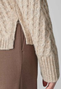 beige Strickpullover mit einem Kabelmuster, gerippten Bündchen und einem seitlichen Schlitz, kombiniert mit braunen Stoffhosen, die strukturierte Materialien präsentieren.