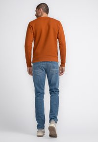 Oranje sweatshirt met textuur en geribbelde manchetjes, gecombineerd met slim-fit blauwe spijkerbroek en lichte sportschoenen. De achterkant toont de details van de outfit.