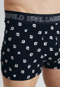 Fekete boxer alsónadrág, amelyen ismétlődő mesehős arcok mintázata található, fehér és bézs részletekkel kiemelve. Rugalmas derékrész, "KARL LAGERFELD" felirattal.