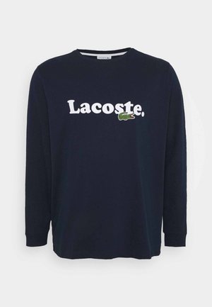 Camiseta de manga larga color azul marino, confeccionada en algodón, con el logo blanco "Lacoste" y un emblema de cocodrilo verde en el lado izquierdo.
