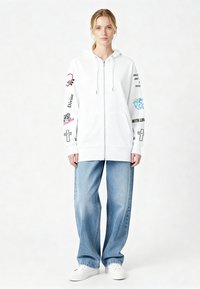 Bela kapucar s pajkano z zadrgo in grafikami na rokavih, kombinirana s svetlo modrimi širokimi kavbojkami in belimi supergami. Ima vrvice za zategovanje in žepe.