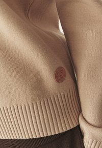 Maglione marrone chiaro con bordo e polsini a coste, realizzato in un tessuto a maglia texturizzato. Include una piccola etichetta rotonda in pelle con un logo a forma di orso.