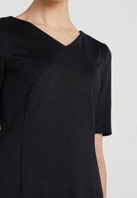 Musta mekko, jossa on V-pääntie ja lyhyet hihat, valmistettu tekstuuripinnasta, jossa on vino sauma yksityiskohta bodysessa.