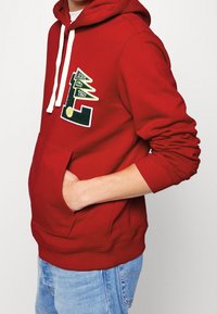 Personne portant un sweat à capuche rouge avec des cordons blancs et un patch Lacoste 1927, mains dans les poches, associé à un jean bleu clair.