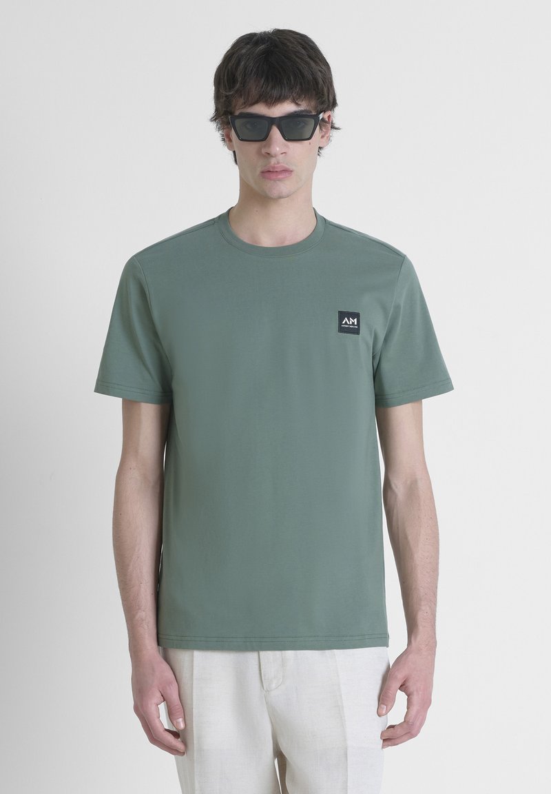 T-shirt in cotone verde a maniche corte con scollo rotondo e una piccola etichetta logo nera sul lato sinistro del petto. Modello che indossa occhiali da sole scuri.