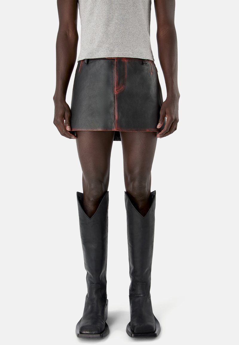 Persona che indossa una minigonna in pelle nera con cuciture rosse, top grigio senza maniche e stivali neri alti con punte quadrate su sfondo bianco.
