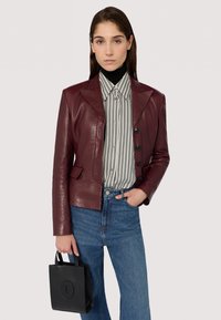 Giacca in pelle bordeaux con una silhouette su misura, caratterizzata da un colletto a punta e bottoni neri, abbinata a una camicia a righe e jeans a vita alta. Una piccola borsa nera completa il look.