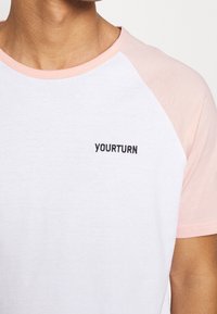 Tričko s krátkým rukávem, bílé barvy s světle růžovými raglánovými rukávy. Černé vyšívané logo "YOURTURN" na hrudi. Měkká bavlněná tkanina.