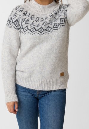 Personne portant un pull tricoté gris clair avec des motifs géométriques noirs sur la poitrine et un jean bleu.
