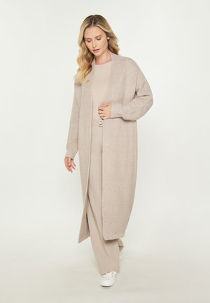 Long, open-front cardigan in light beige, knitted texture. Features wide sleeves and ribbed cuffs, worn over a matching beige outfit and white sneakers.