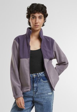 Lila und graue Fleecejacke mit hohem Kragen, Reißverschluss und gerippten Bündchen, im Color-Blocking-Design. Über einem schwarzen Oberteil getragen.