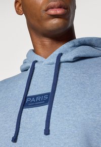 Blå sweatshirt med hætte, udstyret med en marineblå snor og et rektangulært marineblåt mærke med "PARIS SAINT-GERMAIN" i hvid.