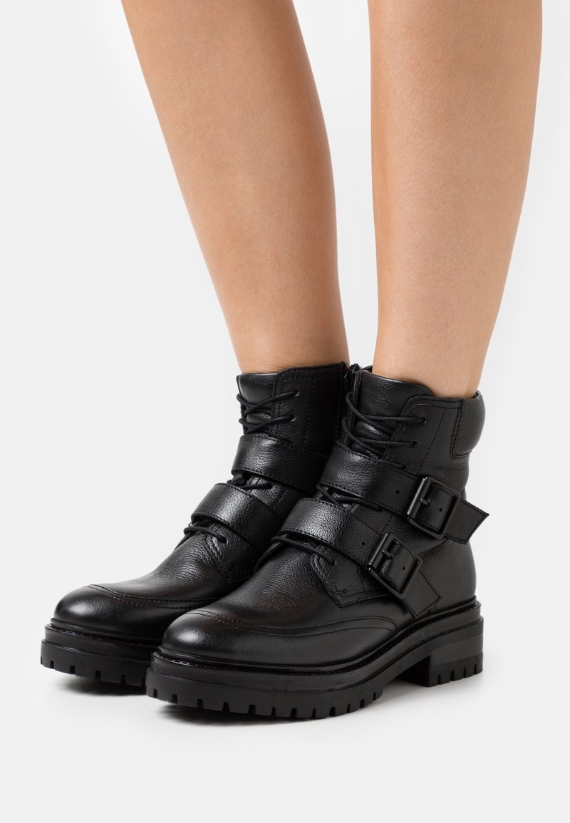 Bottines en cuir noir avec semelles épaisses, boucles, lacets et fermetures éclair latérales portées sur des jambes nues sur fond blanc.