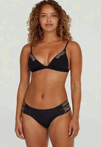 Γυναίκα με σγουρά μαλλιά, φορώντας ένα μαύρο μπικίνι με μπεζ κέντημα σε φύλλο στο πάνω μέρος και στις πλευρές, στέκεται μπροστά σε απλό φόντο.