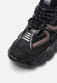 Zapatilla negra que combina texturas brillantes y de malla, con una suela gruesa, un logo destacado y patrones de rayas distintivos.