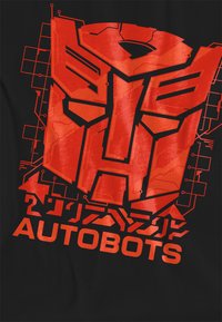 Schwarzes T-Shirt mit einem großen roten Grafik eines Robotersymbols, das komplexe Linienelemente aufweist, und dem Wort "AUTOBOTS" in fetter roter Schrift darunter.