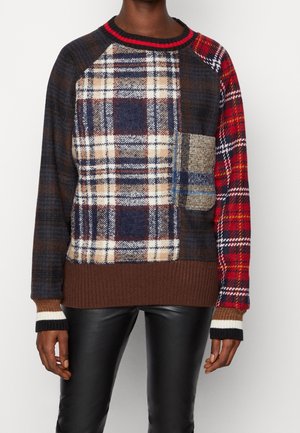 Personne portant un pull patchwork à carreaux multicolores avec une petite poche et un ourlet côtelé marron, associé à un pantalon en cuir noir.