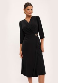 Czarna sukienka wrap z rękawami do trzech czwartych, z dekoltem w kształcie litery V, akcentem w postaci złotej paska oraz spódnicą sięgającą kolan z bocznym rozcięciem.