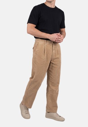 Man draagt een zwart t-shirt, beige ribfluwelen broek en beige sneakers, staat met gevouwen handen voor zich tegen een effen achtergrond.