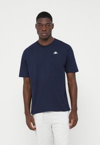 Joven vestido con una camiseta Kappa de color azul marino y pantalones de chándal grises claros, de pie contra un fondo blanco.