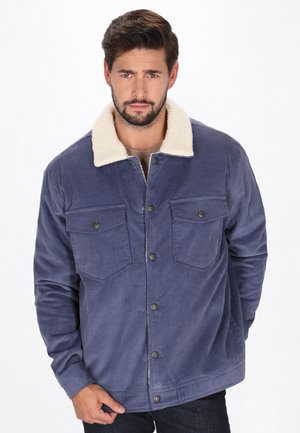 Bărbat purtând o jachetă din corduroy albastră cu guler de fleece alb, buzunare cu nasturi și capse metalice pe fundal alb.