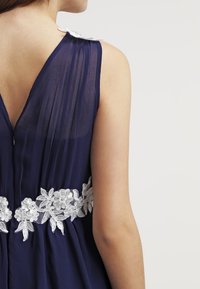 Námořnické modré šaty se síťovanou tkaninou, s komplexní bílou květinovou krajkou v pase a zdobené bílým akcentem na rameni.