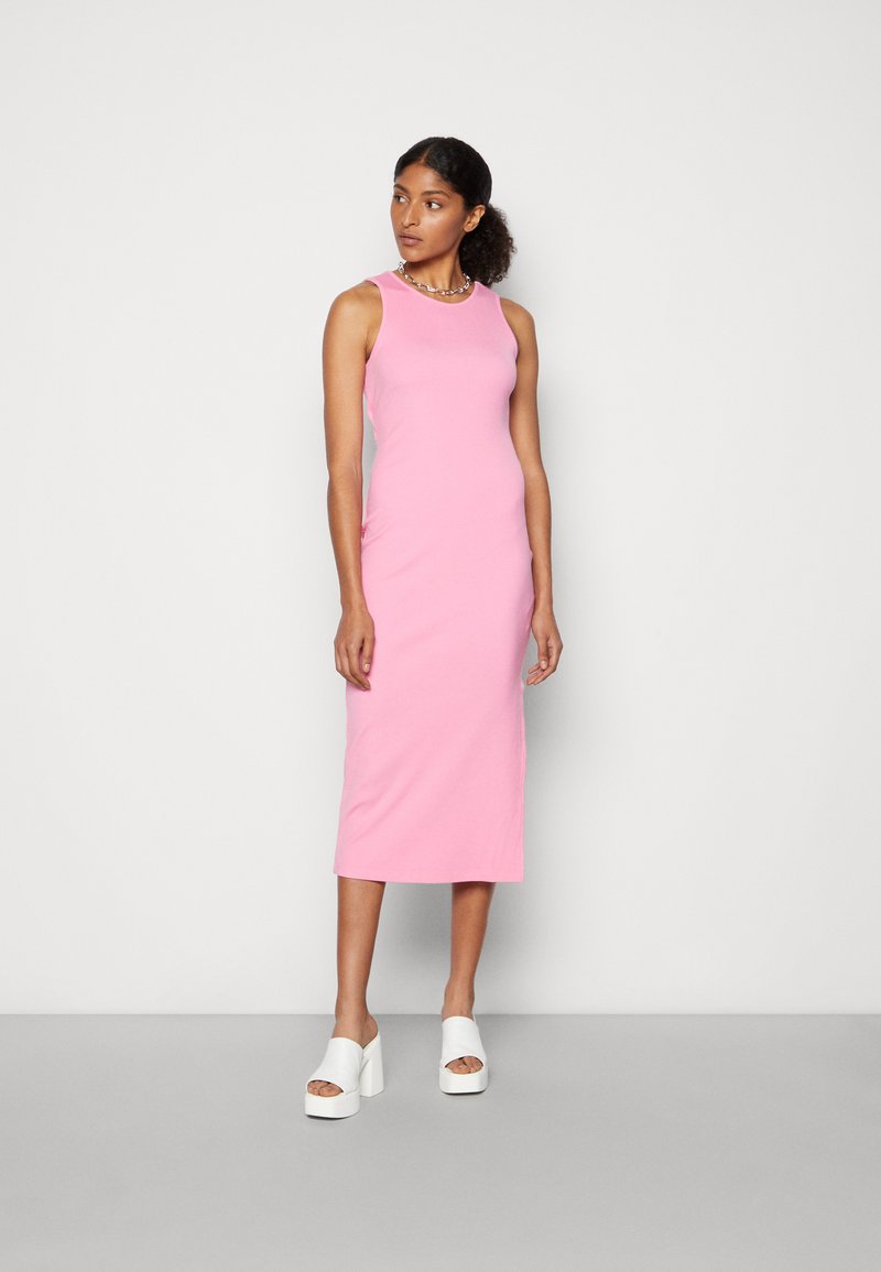 Vestido midi rosa sem mangas, feito de algodão suave, apresentando uma silhueta ajustada. Combinado com sapatos de plataforma brancos de bico aberto.