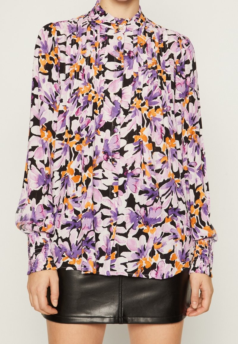 Femme portant un chemisier à manches longues à motif floral violet, orange et noir, associé à une mini-jupe en cuir noire.