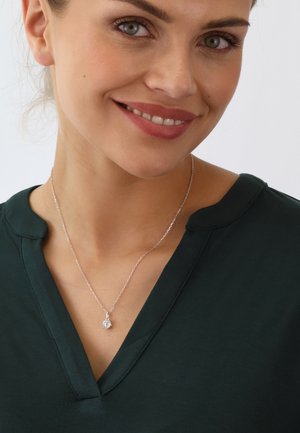 Silberne Halskette mit einem runden Anhänger, getragen von einer Person in einem dunkelgrünen Shirt mit V-Ausschnitt. Glattes Material und polierte Metallelemente.