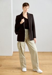 Schwarze maßgeschneiderte Blazer über einem schwarzen gerippten Oberteil, kombiniert mit lockeren hellbeigen Cargo-Hosen mit Reißverschlüssen und Taschen sowie weißen Sneakers.