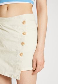 Jupe portefeuille en coton beige clair présentant une coupe diagonale et cinq boutons ronds de couleur claire sur le devant, avec un détail d'ourlet.