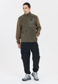 Fleece jacket in olive green with a zip closure, zippered pocket, and textured sleeves, paired with black cargo pants and grey hiking boots.