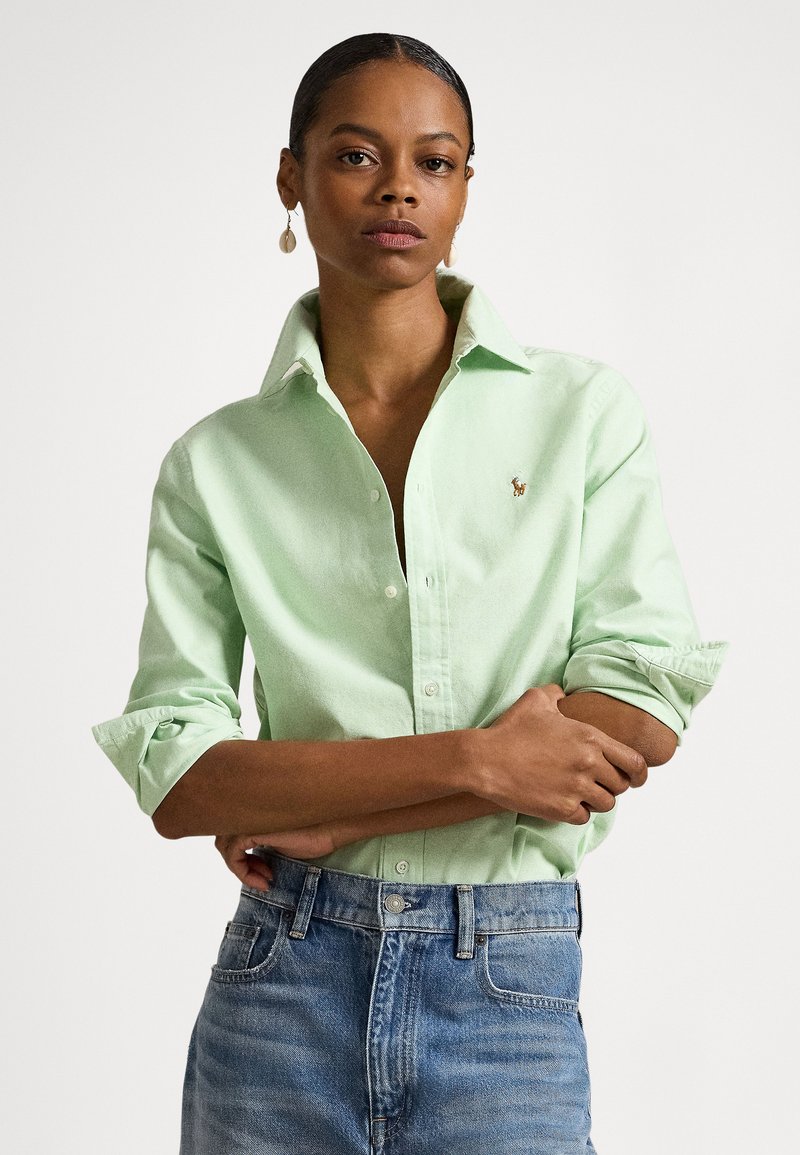 Gaiši zaļa pogām aiztaisāma krekls ar salocītām piedurknēm, uz krūtīm neliela izšūta logo, kopā ar klasiskām zilām džinsa biksēm.