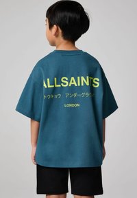 Teal bomulds T-shirt med korte ærmer, der har store gule bogstaver "ALLSAINTS" og japanske tegn på ryggen. Sorte shorts bæres.