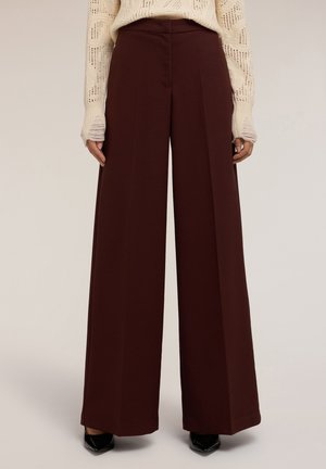 Mujer vestida con pantalones anchos de color marrón oscuro, suéter de punto crema con agujeros para los pulgares y zapatos negros de punta, de pie frente a un fondo liso.