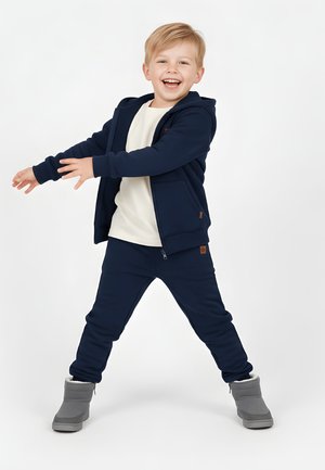 Uśmiechnięty młody chłopiec w granatowej bluzie z kapturem i spodniach, białej koszulce oraz szarych zimowych butach stoi z wyciągniętymi ramionami i nogami rozstawionymi na białym tle.