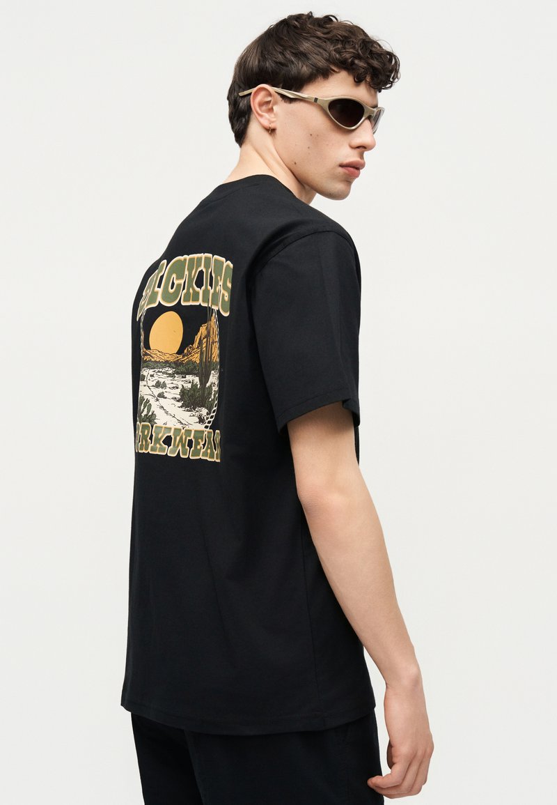 Giovane uomo con occhiali da sole e una maglietta nera con grafica di un paesaggio desertico e sole sul retro, di profilo su uno sfondo semplice.