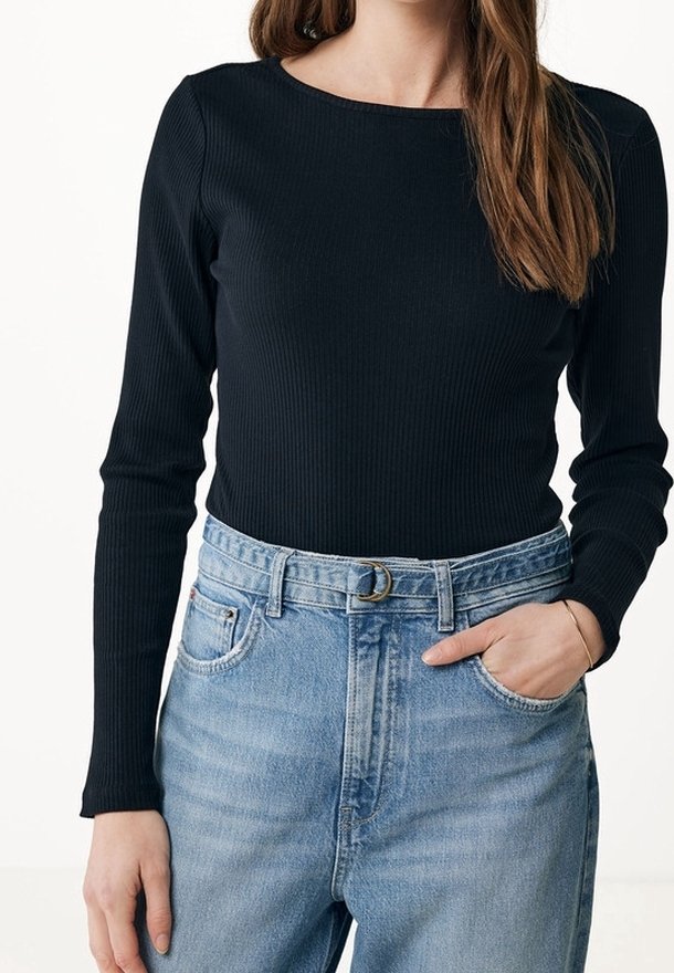 Femme portant un haut noir côtelé à manches longues, rentré dans un jean taille haute bleu clair avec une ceinture et la main dans la poche.