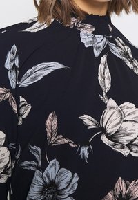 Schwarze Blumenbluse mit hohem Ausschnitt, die sanfte rosa und blaue Blumenmuster auf einem dunklen Hintergrund und aus strukturiertem Stoff zeigt.