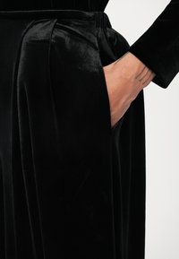 Fekete bársony nadrág pliszé részletezéssel, oldalzsebbe és puha, sima textúrába illeszkedve. Közeli nézet, kéz a zsebben.