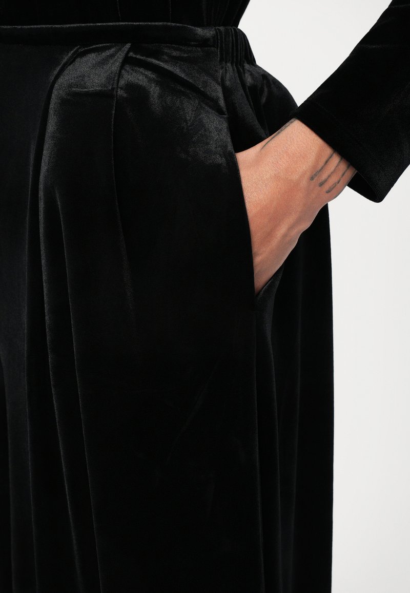 Black velvet pants with pleated detailing, featuring a side pocket and a soft, smooth texture. Close-up view of hand in pocket.