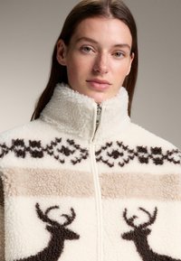 Kurtka polarowa w kolorze kości słoniowej z brązowymi wzorami jelenia. Posiada wysoki kołnierz i zamek błyskawiczny oraz dekoracyjne poziome paski w beżu.