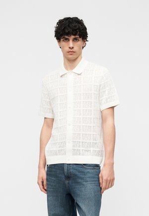 Junger Mann mit lockigem schwarzem Haar, der ein weißes kurzärmeliges Spitzen-Strickhemd mit Knöpfen und eine blaue Jeans trägt, vor einem schlichten Hintergrund stehend.