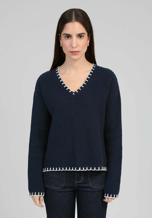 Femme aux longs cheveux foncés portant un pull en maille bleu marine avec des bordures blanches et un jean foncé, vue de face sur un fond gris uni.