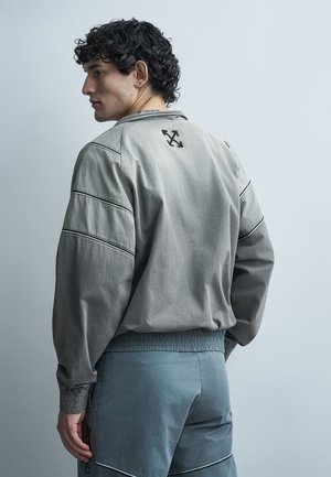 Άνδρας με σγουρά μαλλιά που φοράει γκρι μπουφάν με λογότυπο στην πλάτη και ταιριαστό γκρι παντελόνι, στραμμένος προς τα αριστερά μπροστά σε απλό φωτεινό φόντο.