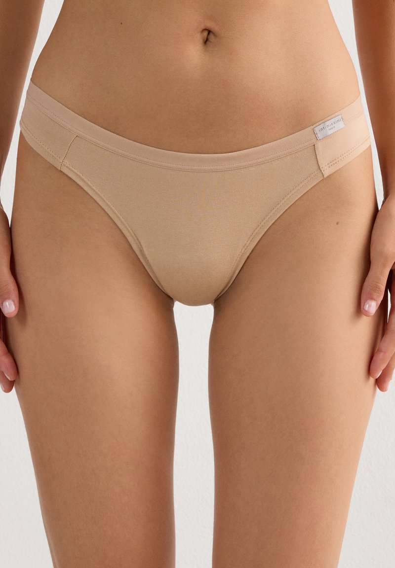 Žena nosící béžové bezešvé bikiny s nízkým pasem, stojící s rukama volně podél těla na jednobarevném pozadí.