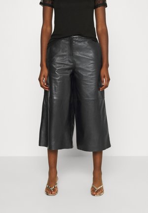 Woman wearing black leather wide-leg cropped pants, black top with lace sleeves, and snakeskin-patterned sandals, standing against white background.