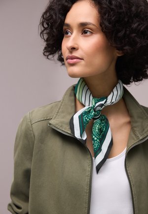 Femme aux cheveux courts et bouclés portant une veste vert olive et une écharpe à motifs verts, blancs et noirs nouée autour du cou.