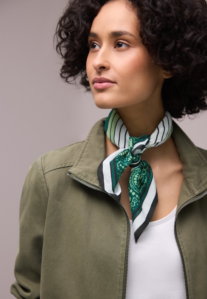 Kobieta o krótkich, kręconych włosach, ubrana w oliwkową kurtkę i zielono-biało-czarny szal we wzory, zawiązany wokół szyi.