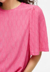 Top rosa testurizzato con un motivo a onde, caratterizzato da maniche corte e ampie e da una leggera arricciatura alle spalle. Finitura del tessuto liscia.